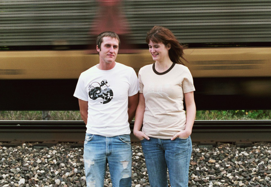 Photograph of a man and woman modeling graphic t-shirts while standing in front of a passing train moving at high speed.