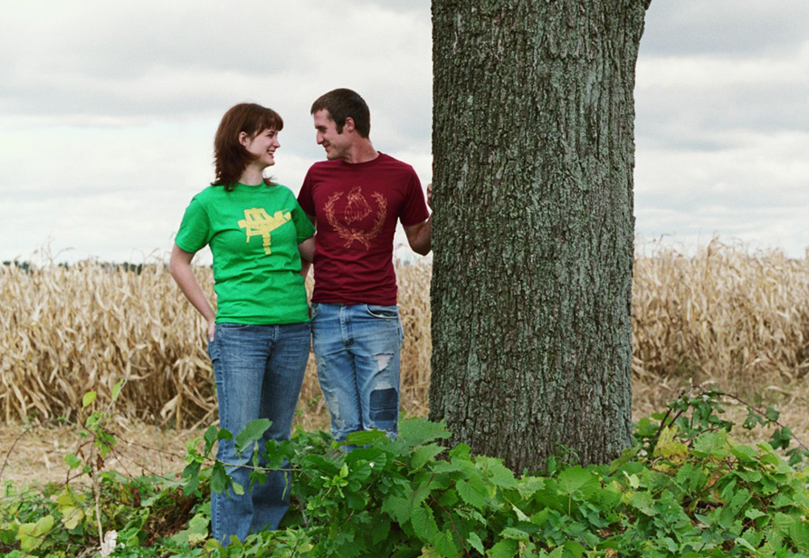 Photograph of a man and woman looking at each other lovingly while standing next to a tree and modeling graphic t-shirts in front of a corn field.