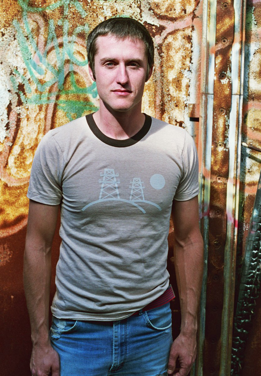 Photograph of a man modeling a beige t-shirt with a light blue geometric illustration depicting hills and electricity pylons.