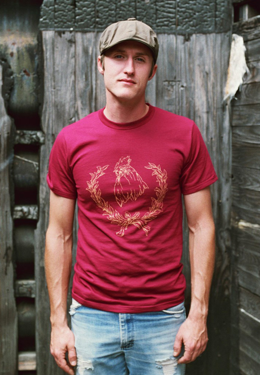 Photograph of a man modeling a red t-shirt with a gold illustration of a rooster and laurels.