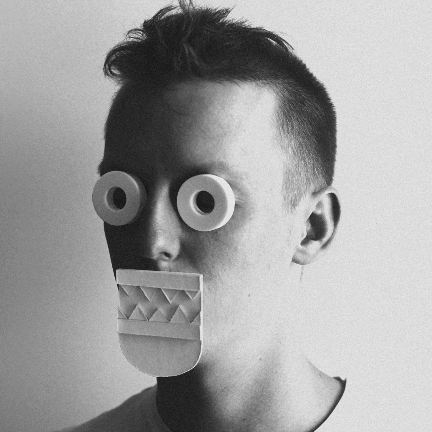 A black and white photograph with a three quarter view of a young man's face wearing gometric paper shapes covering his eyes and mouth.